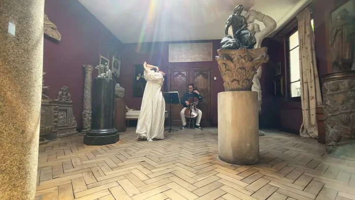 La danseuse en robe blanche, bras levés, entre les sculptures d’une salle pourpre